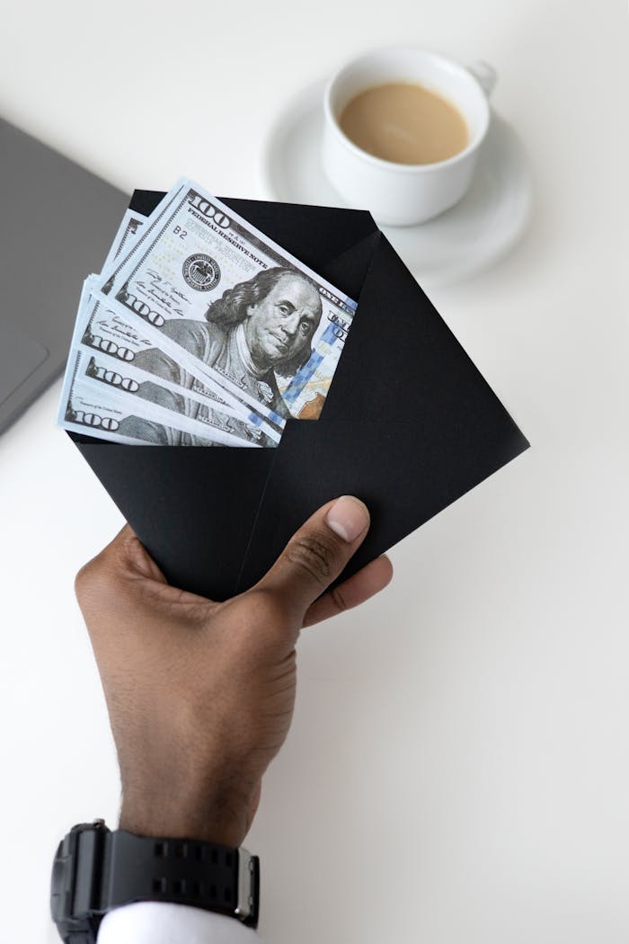 Close-up of a hand holding an envelope with dollar bills, next to a coffee cup.